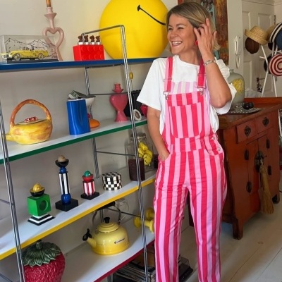 Pessoa com macacão rosa e branco às riscas e t-shirt branca junto a estante colorida com objetos decorativos