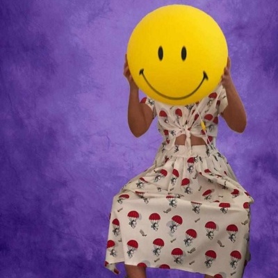 Vestido branco com padrão de paraquedas vermelho, pessoa segura bola amarela sorridente, fundo roxo.