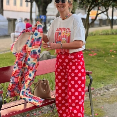 Pessoa com t-shirt branca APEROL Spritz e calças de bolinhas vermelhas a segurar roupa colorida junto a banco vermelho num parque.