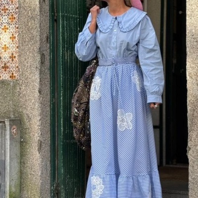 Mulher com vestido comprido azul com gola grande e saia riscada azul com branco e rendas brancas, óculos escuros, bolsa marrom, junto a porta verde e parede de pedra