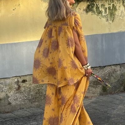 Mulher de costas com conjunto amarelo estampado, segurando guarda-chuva, junto a muro amarelo e cinzento.