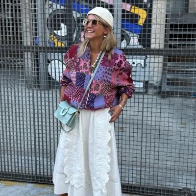 Mulher com blusa patchwork colorida, saia branca longa, chapéu de malha branco, sapatilhas rosa e bolsa azul clara em frente a grade metálica com graffiti.