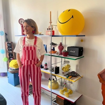 Macacão às riscas rosa e vermelho com t-shirt branca em sala com prateleira colorida e objetos decorativos