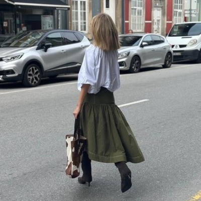 Mulher de costas na rua com blusa branca, saia verde e bolsa castanha e branca