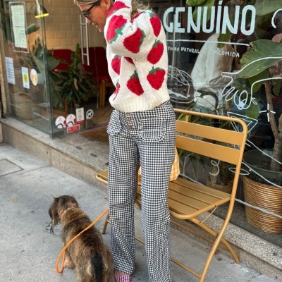Mulher vestindo roupa com padrão de morangos e calças xadrez, levando um cão com uma trela laranja numa rua junto a uma cadeira amarela diante de uma loja