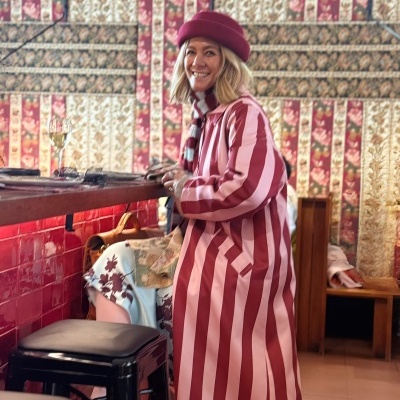 Mulher com casaco riscas vermelho e rosa sentada em banco alto em espaço interior decorado com azulejos vermelhos e parede floral.