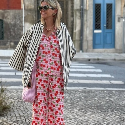 Mulher com pijama estampado de cerejas, casaco riscado preto e branco, óculos de sol, bolsa rosa, em rua com calçada de pedras e porta azul