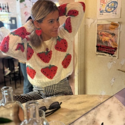 Mulher vestindo camisola branca com morangos vermelhos e brincos de coração vermelhos sentada num café.