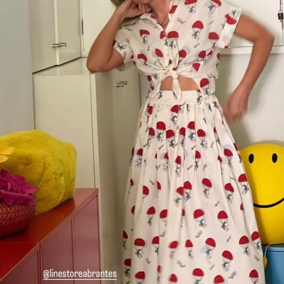 Mulher veste conjunto branco com padrão de guarda-chuvas vermelhos e figuras pequenas pretas, com nó frontal na camisa.