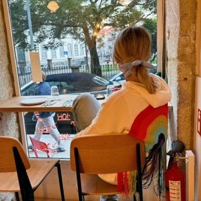 Pessoa sentada com hoodie branco com arco-íris colorido nas costas junto a janela e mesa de madeira