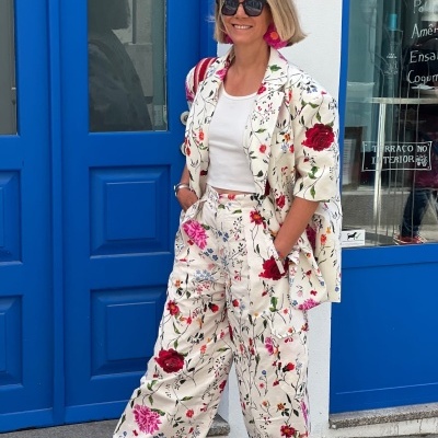 Mulher com conjunto floral creme vermelho e rosa, brincos grandes, sandálias vermelhas, porta azul e janela com texto em fundo urbano.