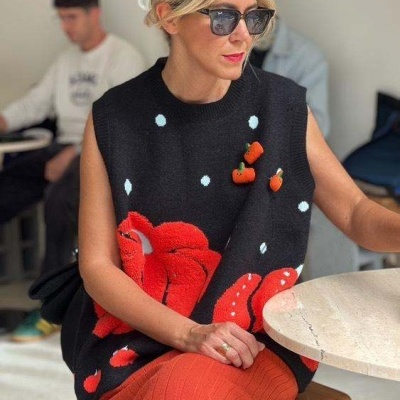 Colete preto com flores vermelhas e bolinhas azuis, saia laranja, mulher com óculos de sol sentada à mesa