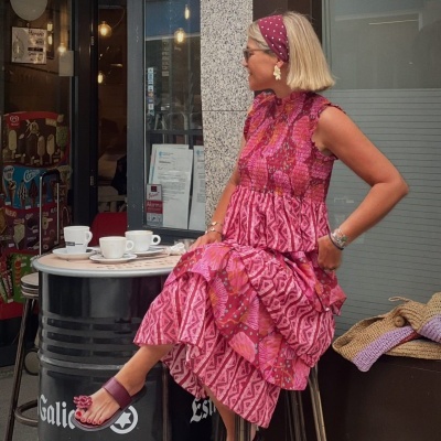 Mulher com vestido rosa estampado em banco metálico junto a mesa redonda com cafés.