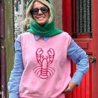 Mulher com camisola rosa de malha com lagosta vermelha, camisa azul riscada, lenço verde e óculos escuros na rua.