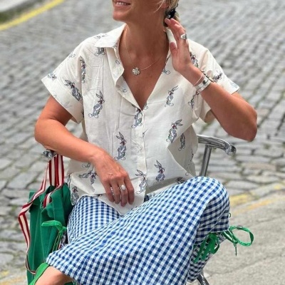 Mulher com camisa estampada de coelhos, calças xadrez azuis e brancas e sapatos verdes sentada ao ar livre.