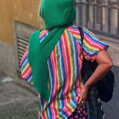 Pessoa com camisola de riscas coloridas, cachecol verde, calças azuis e mochila preta na rua