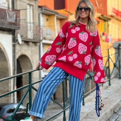 Mulher com suéter vermelho de morangos e cerejas, calças às riscas azuis, óculos escuros e sapatos brancos a sorrir numa rua urbana.