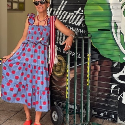 Mulher com vestido azul de bolinhas vermelhas e chapéu de palha ao lado de carrinho verde numa rua.