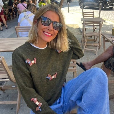 Mulher com suéter verde com cães de tecido, calças azuis e óculos Ray-Ban numa esplanada.