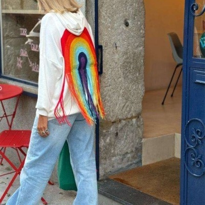 Mulher com suéter branco com capuz e arco-íris em lã colorida nas costas, calças azuis largas, perto de porta azul e móveis vermelhos na rua