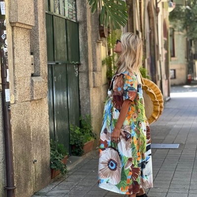 Mulher vestindo vestido longo estampado e mochila redonda de palha numa rua de calçada com edifícios antigos