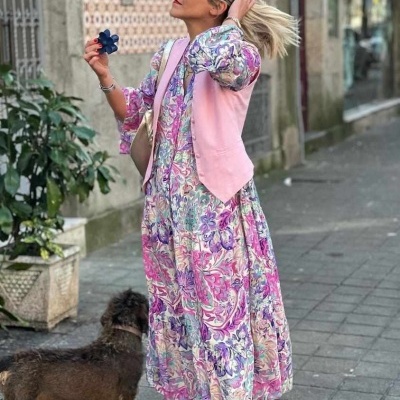 Mulher vestindo vestido floral e colete rosa com cão ao lado numa calçada urbana