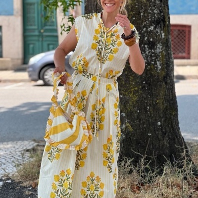 Mulher com vestido floral amarelo e bolsa riscada amarelo e branca junto a árvore ao ar livre