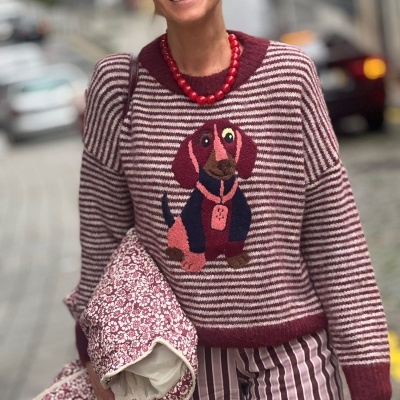 Pessoa com camisola de malha listrada bordeaux e bege com bordado de cachorro colorido, calças listradas, colar vermelho e óculos de sol.