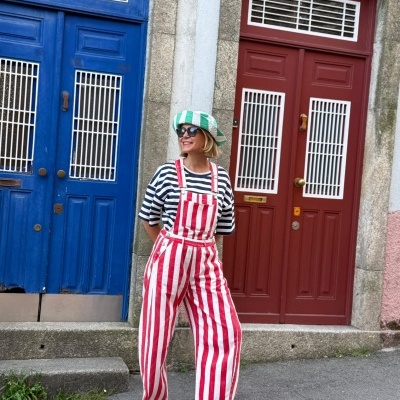 Pessoa com macacão riscas vermelhas e brancas, tshirt riscas pretas e brancas, chapéu verde e branco, chinelos amarelos, junto a portas azuis e vermelhas