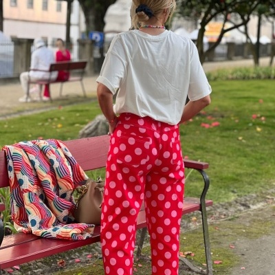 Mulher com calças vermelhas de bolinhas brancas num parque junto a banco vermelho com casaco colorido e mala bege