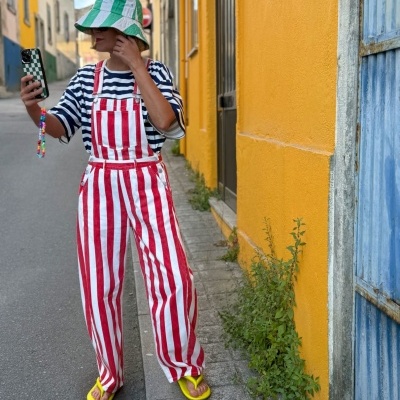Pessoa com roupa às riscas, chapéu verde e branco, chinelos amarelos, a tirar selfie na rua com paredes coloridas amarelas e azuis.