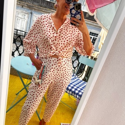 Mulher com conjunto branco e vermelho estampado, sapatos vermelhos e óculos escuros numa varanda com cadeira e mesa coloridas