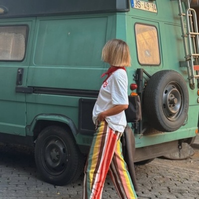 Pessoa com t-shirt branca e calças às riscas coloridas ao lado de veículo verde antigo na calçada