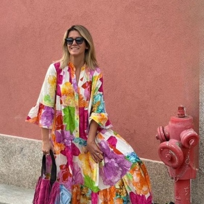 Mulher com vestido floral colorido, óculos escuros, calçado desportivo verde e branco, segura malas rosa e azul junto a hidrante vermelho e parede rosa.