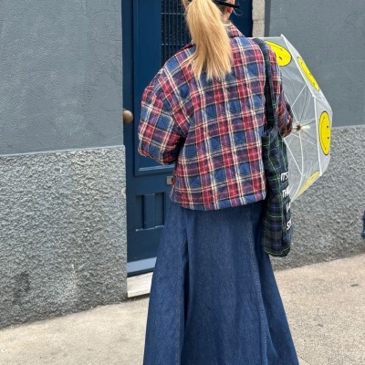 Mulher com casaco xadrez, saia comprida azul, guarda-chuva transparente com carinhas amarelas, boné azul, em frente a porta azul e parede cinza