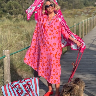 Mulher com vestido rosa com flores laranja e túnica rosa com flores brancas segurando túnica, bolsa riscada azul e vermelha ao lado e cão com trela vermelha