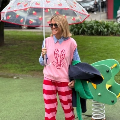 Mulher com colete rosa com lagosta, calças riscas e guarda-chuva transparente com morangos