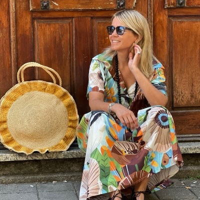 Mulher loira com vestido colorido sentada junto a porta de madeira com bolsa de palha redonda.