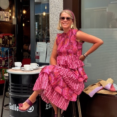Mulher com vestido rosa estampado e sandálias vinhos sentada numa esplanada com três chávenas de café em balcão preto.