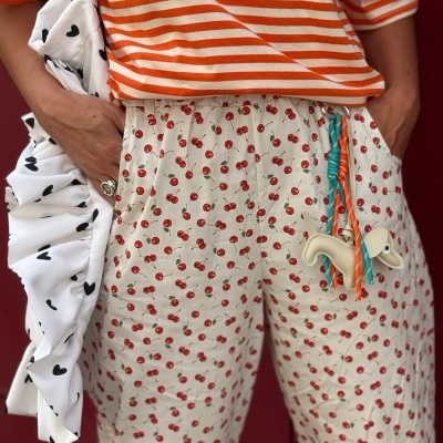 Calças bege com padrão de cerejas vermelhas, cordão colorido e chaveiro, t-shirt às riscas laranja e branca, bolsa branca com corações pretos.