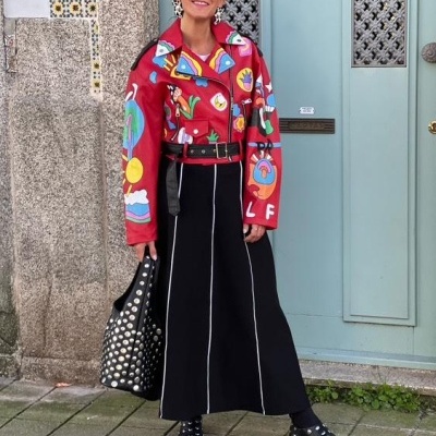 Mulher com casaco vermelho estampado, saia preta, botas e bolsa com tachas, em frente a porta azul.