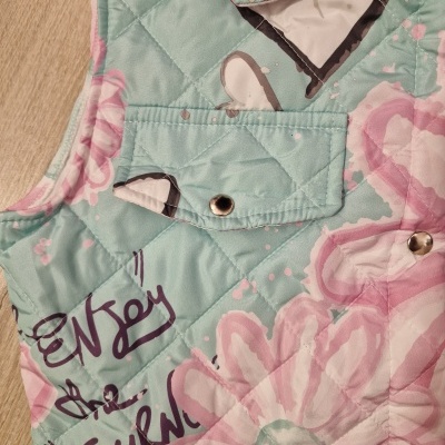 Colete acolchoado verde água com flores rosa e texto manuscrito em preto.