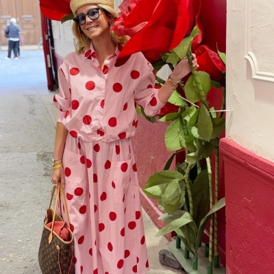 Mulher com vestido rosa de bolinhas vermelhas e chapéu de palha segura flor vermelha gigante junto a parede vermelha