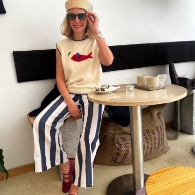 Mulher com calças às riscas, camisola com peixe, chapéu de palha e óculos escuros sentada num café.