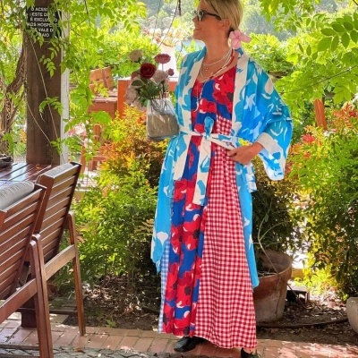 Mulher com vestido longo colorido floral e quimono azul claro segurando vaso com flores ao ar livre