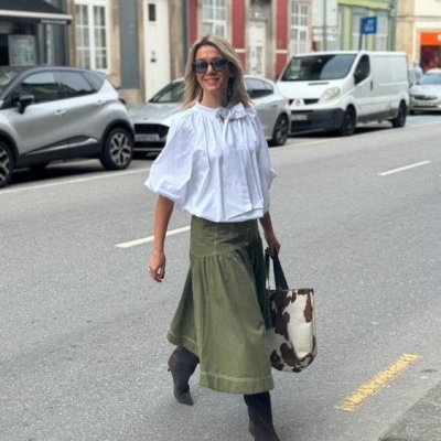 Mulher com blusa branca e saia verde militar caminhando na rua com bolsa e botas