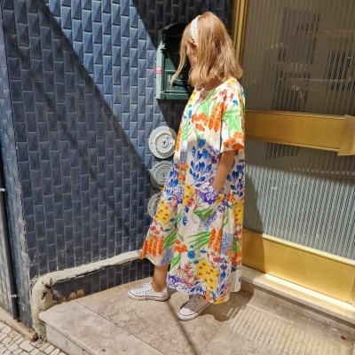 Mulher com vestido floral colorido e ténis brancos diante de porta dourada e parede azul