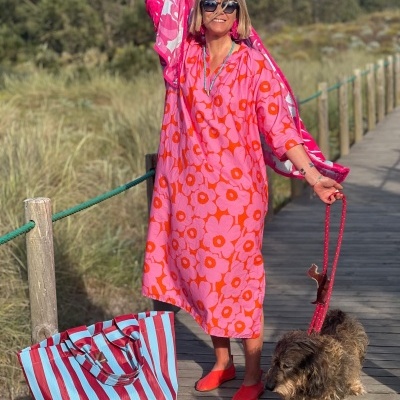 Mulher com vestido rosa de flores vermelhas, mala às riscas azul e vermelha, e cão castanho num passadiço de madeira.