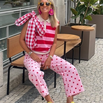 Mulher sentada com roupa rosa, vermelha e amarela, óculos de sol e banco numa calçada.