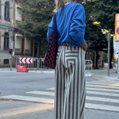 Mulher com pulôver azul, calças às riscas, mala xadrez azul e laranja numa rua urbana